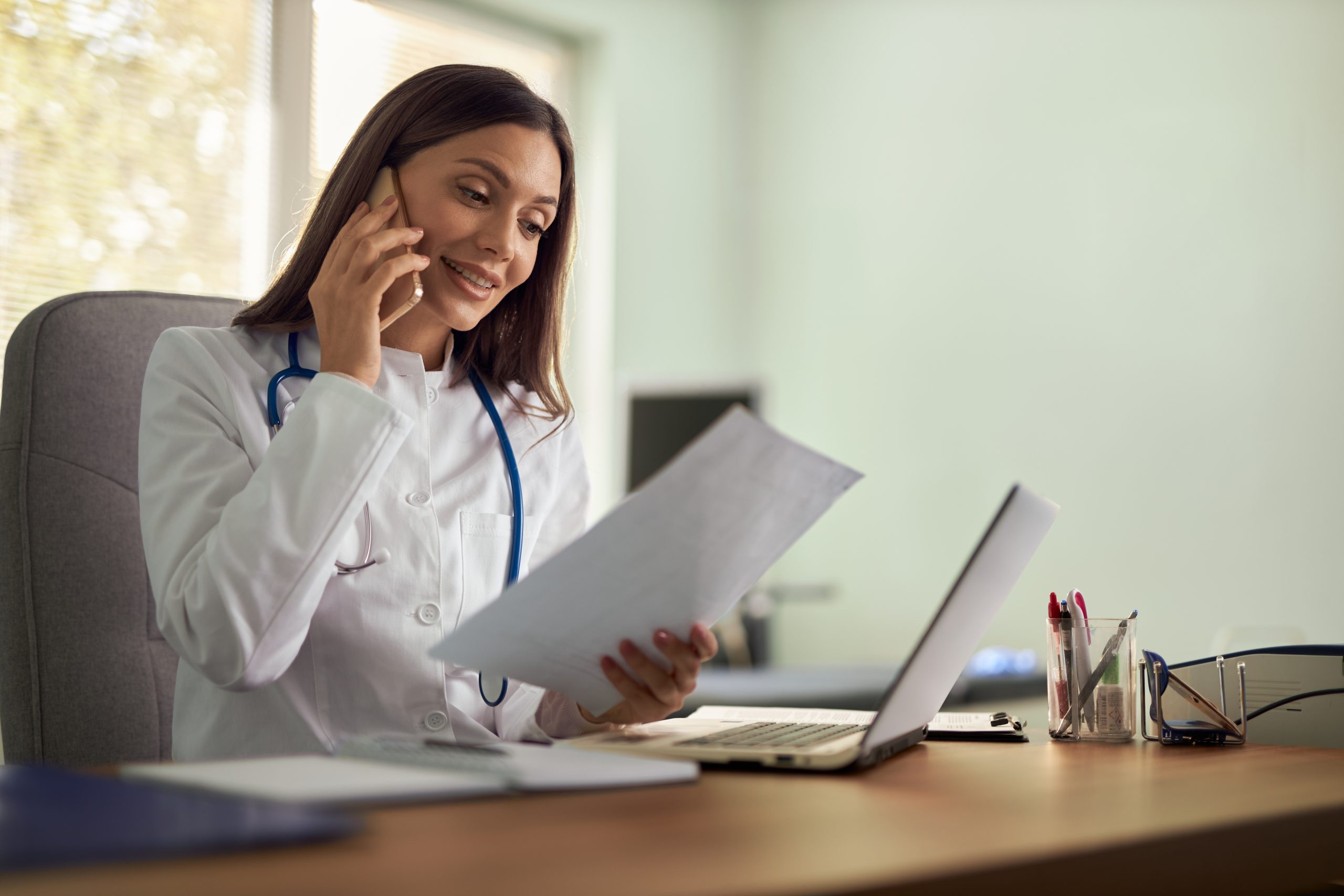 Young female doctor reading medical documents in her office while talking on cell phone.